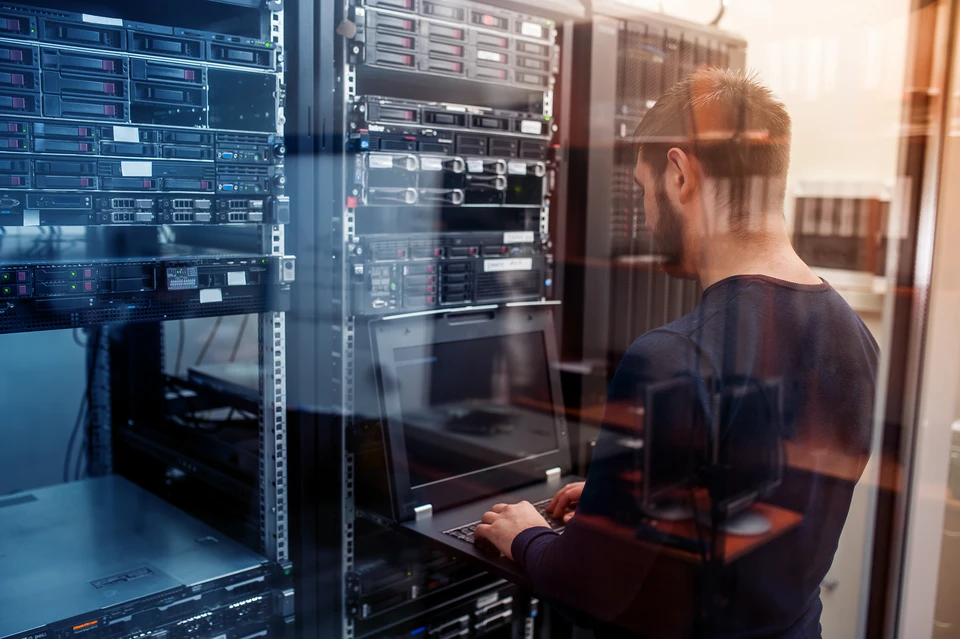 Un technicien informatique travaille dans une salle serveur, utilisant un ordinateur portable pour administrer l'infrastructure
